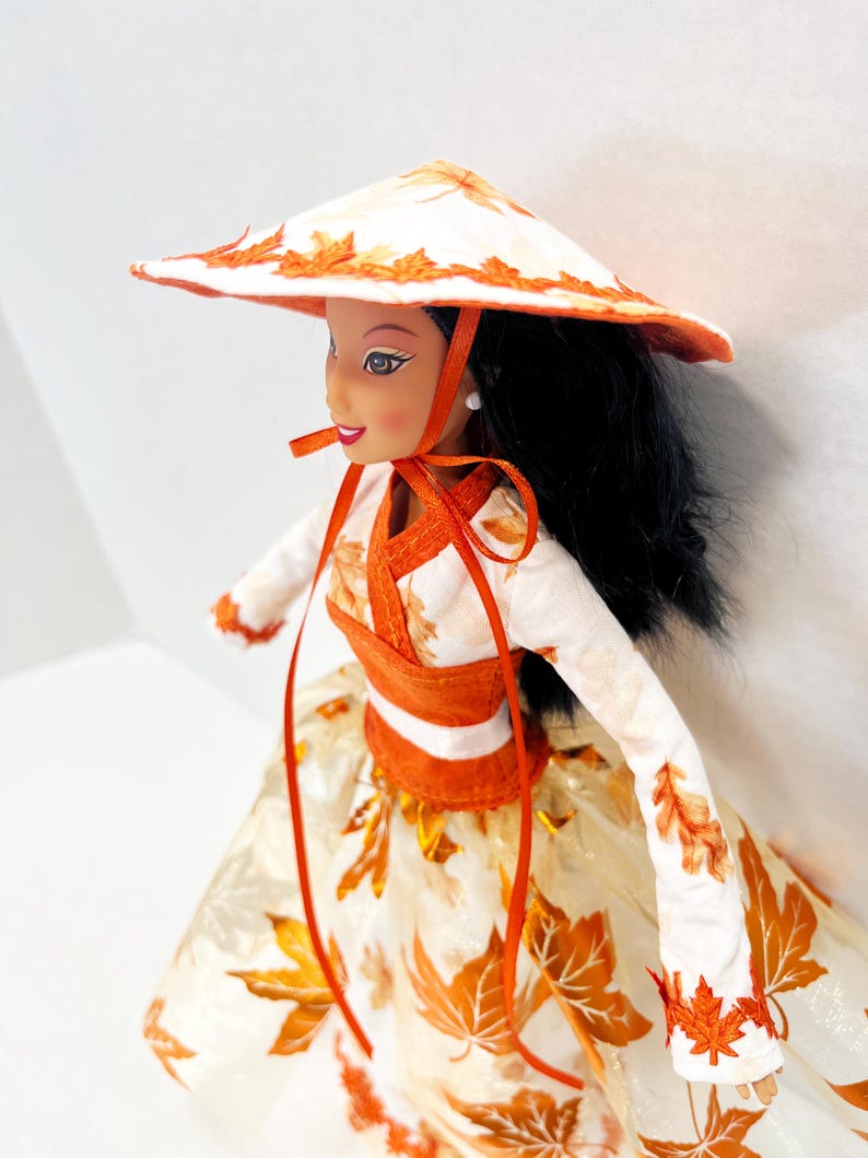 May include: A doll with long black hair wearing a white conical hat and a long-sleeved top featuring orange leaf patterns. The outfit includes a matching full skirt decorated with orange maple leaf prints and orange ribbon ties under the chin.