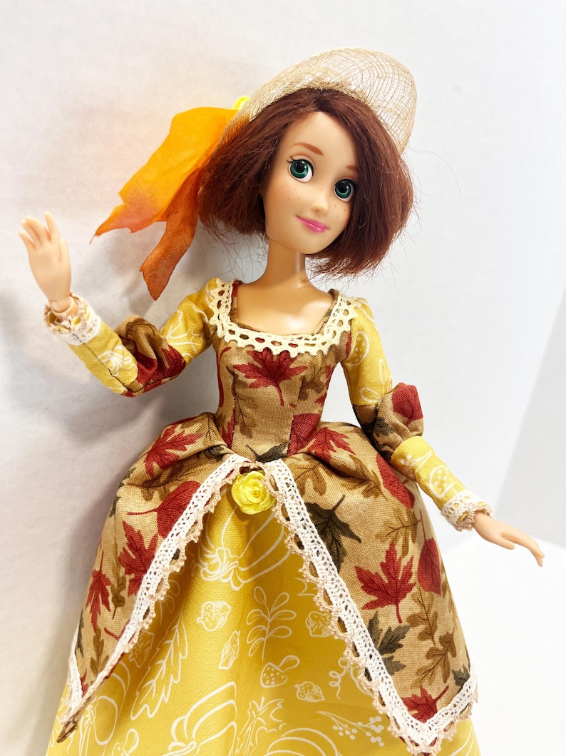 May include: A doll with shoulder-length auburn hair wearing a tan hat with an orange ribbon. The doll wears a dress featuring a brown and red maple leaf print on the bodice and sleeves, with a yellow skirt printed with white autumn motifs.