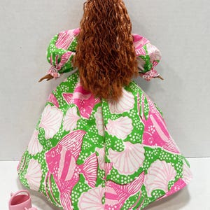 May include: A doll wearing a dress with a pink, green, and white fish and seashell pattern. The doll has long, curly auburn hair and a pink flower in her hair. A small pink watering can is next to the doll.