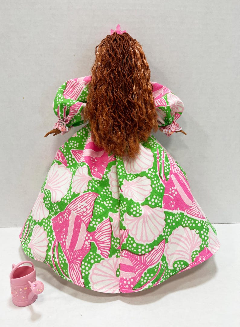 May include: A doll wearing a dress with a pink, green, and white fish and seashell pattern. The doll has long, curly auburn hair and a pink flower in her hair. A small pink watering can is next to the doll.