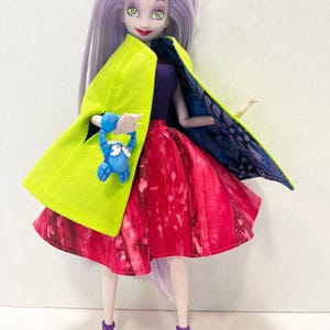 Pu&ograve; includere: Una bambola con lunghi capelli color lavanda, occhi verdi e rossetto rosso. La bambola indossa un corpetto viola, una gonna con motivo rosso e rosa e sandali viola con tacco. Indossa un mantello verde lime con una fodera blu e viola.