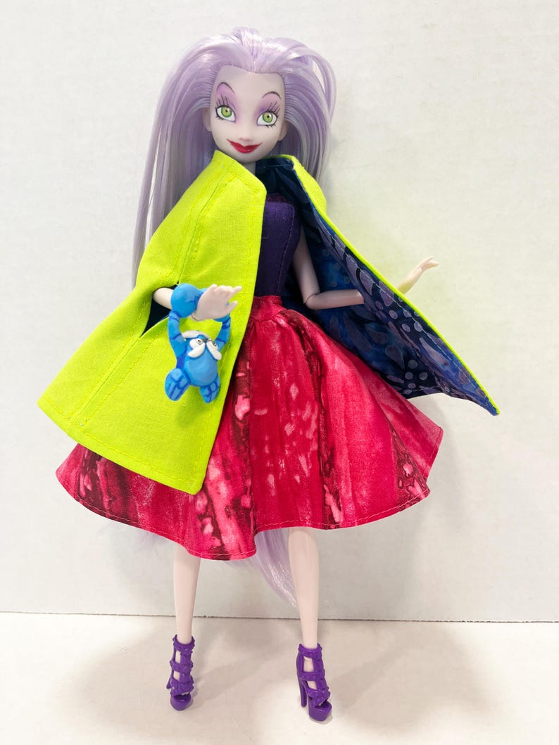 Pu&ograve; includere: Una bambola con lunghi capelli color lavanda, occhi verdi e rossetto rosso. La bambola indossa un corpetto viola, una gonna con motivo rosso e rosa e sandali viola con tacco. Indossa un mantello verde lime con una fodera blu e viola.
