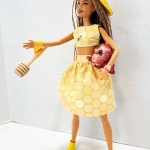 Puede incluir: Mu&ntilde;eca vestida con un conjunto amarillo con estampado de panal, que incluye un top corto y una falda. La mu&ntilde;eca tambi&eacute;n tiene un sombrero amarillo, botas amarillas con suelas rosas y un cuchar&oacute;n de miel de madera. Una criatura rosa se sostiene en los brazos de la mu&ntilde;eca.