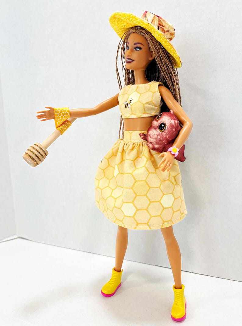 Puede incluir: Mu&ntilde;eca vestida con un conjunto amarillo con estampado de panal, que incluye un top corto y una falda. La mu&ntilde;eca tambi&eacute;n tiene un sombrero amarillo, botas amarillas con suelas rosas y un cuchar&oacute;n de miel de madera. Una criatura rosa se sostiene en los brazos de la mu&ntilde;eca.