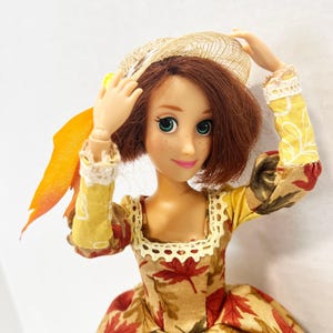 May include: A doll with shoulder-length brown hair and green eyes wearing a long-sleeved dress featuring a pattern of red and brown autumn leaves on a tan background, accented with cream-colored lace trim, a yellow rose, and a straw-textured hat.