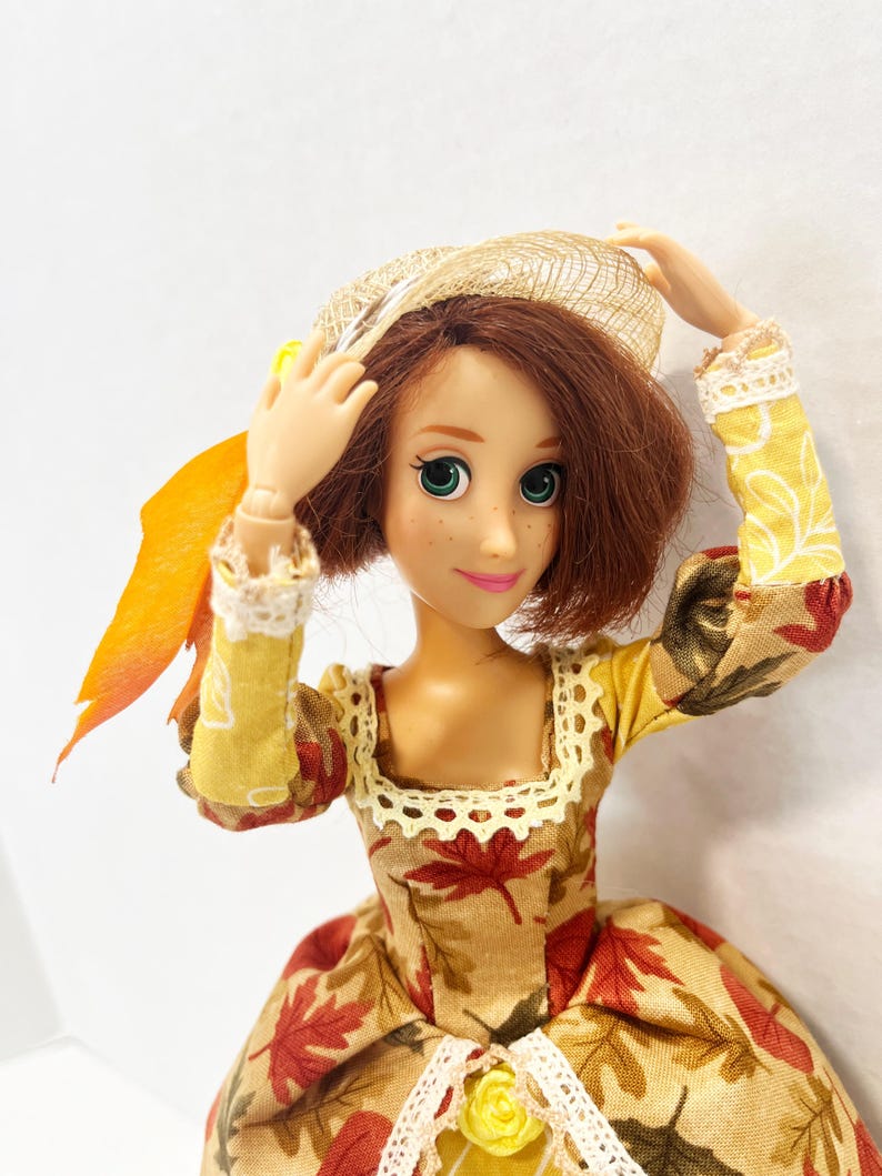 May include: A doll with shoulder-length brown hair and green eyes wearing a long-sleeved dress featuring a pattern of red and brown autumn leaves on a tan background, accented with cream-colored lace trim, a yellow rose, and a straw-textured hat.