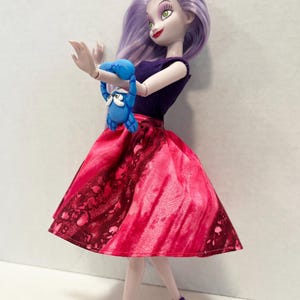 Pu&ograve; includere: Una bambola con capelli color lavanda, occhi verdi e rossetto rosso, che indossa un top viola scuro senza maniche e una gonna rosa vivace con un motivo maculato rosso scuro. La bambola tiene in mano una piccola figura di granchio blu e sandali viola.