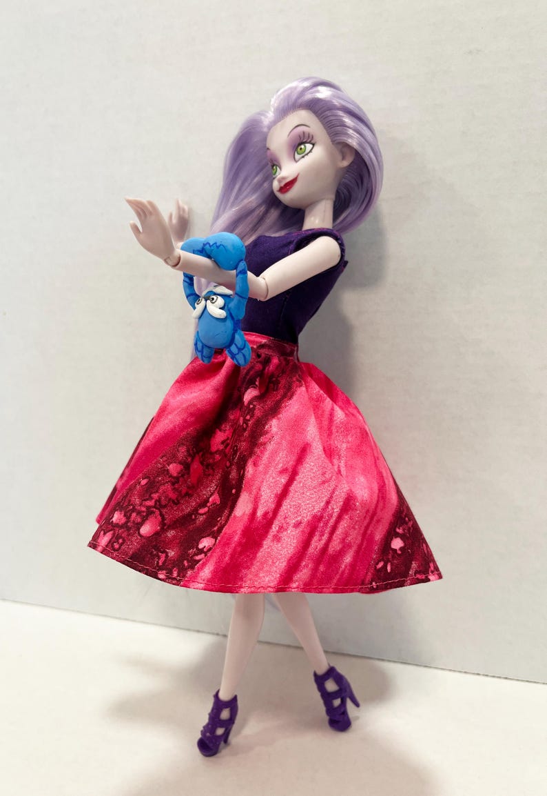 Pu&ograve; includere: Una bambola con capelli color lavanda, occhi verdi e rossetto rosso, che indossa un top viola scuro senza maniche e una gonna rosa vivace con un motivo maculato rosso scuro. La bambola tiene in mano una piccola figura di granchio blu e sandali viola.