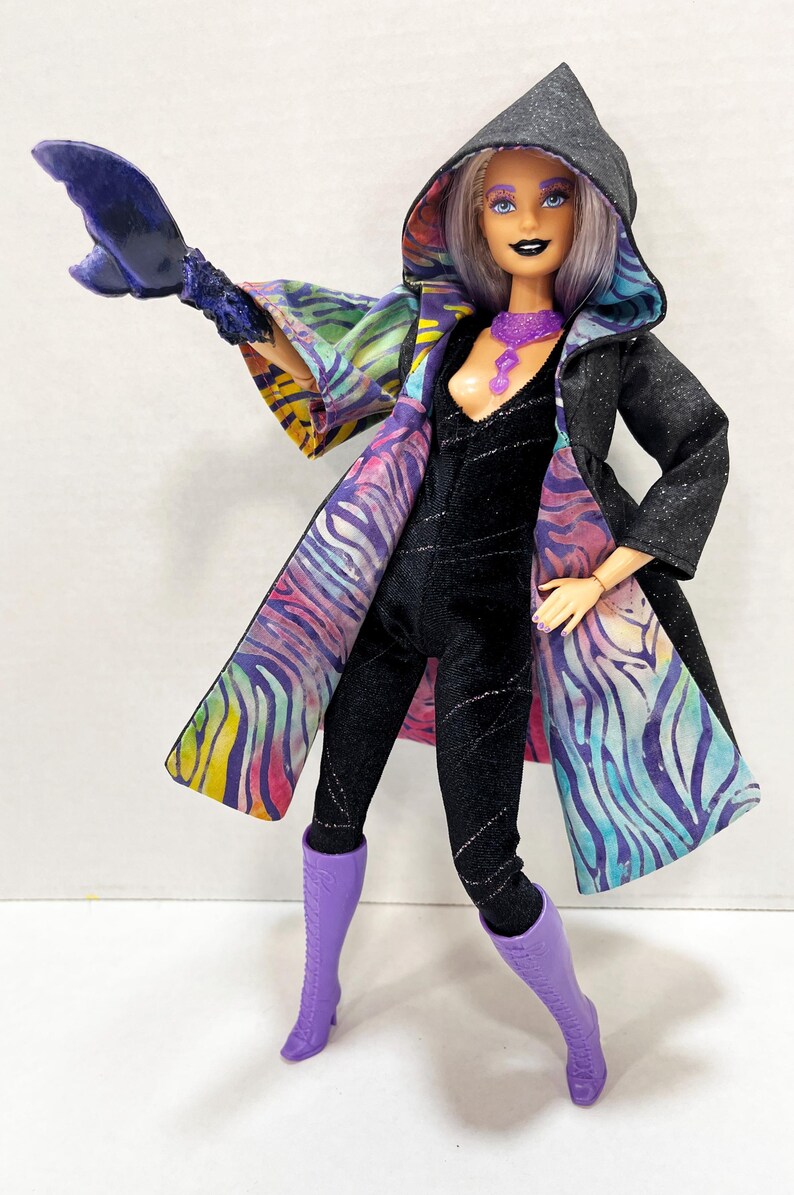 May include: A fashion doll in a black jumpsuit and a hooded coat with a colorful zebra print lining. The doll has purple boots, a purple necklace, and a matching arm accessory. The doll's hair is light purple, and it has dark makeup.