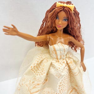 Puede incluir: Una mu&ntilde;eca con piel morena, cabello largo casta&ntilde;o trenzado y una diadema amarilla. La mu&ntilde;eca viste un vestido crema sin tirantes con un estampado de conchas marinas y una superposici&oacute;n transparente. El brazo de la mu&ntilde;eca est&aacute; extendido.