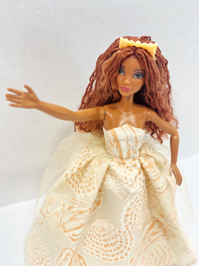 Puede incluir: Una mu&ntilde;eca con piel morena, cabello largo casta&ntilde;o trenzado y una diadema amarilla. La mu&ntilde;eca viste un vestido crema sin tirantes con un estampado de conchas marinas y una superposici&oacute;n transparente. El brazo de la mu&ntilde;eca est&aacute; extendido.
