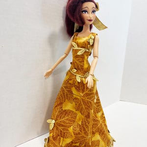 May include: A doll with dark reddish-brown hair styled in a high ponytail, wearing a floor-length mustard yellow gown featuring a brown leaf pattern. The dress is accented with metallic gold leaf-shaped trim along the neckline, waist, and hem.