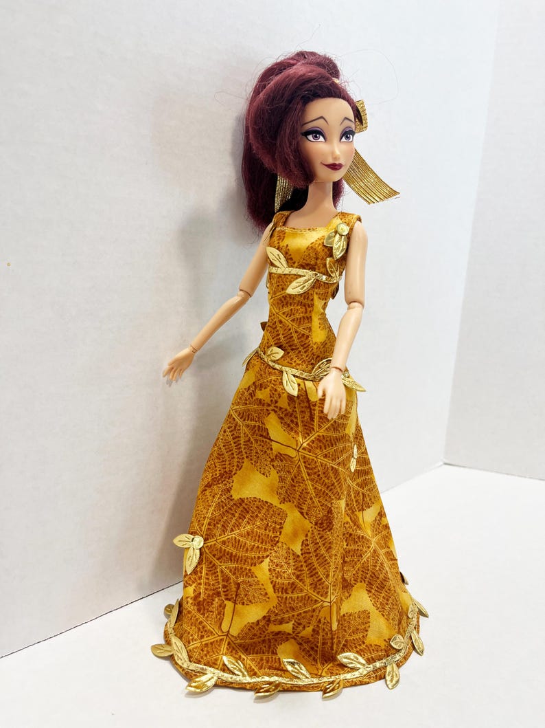May include: A doll with dark reddish-brown hair styled in a high ponytail, wearing a floor-length mustard yellow gown featuring a brown leaf pattern. The dress is accented with metallic gold leaf-shaped trim along the neckline, waist, and hem.