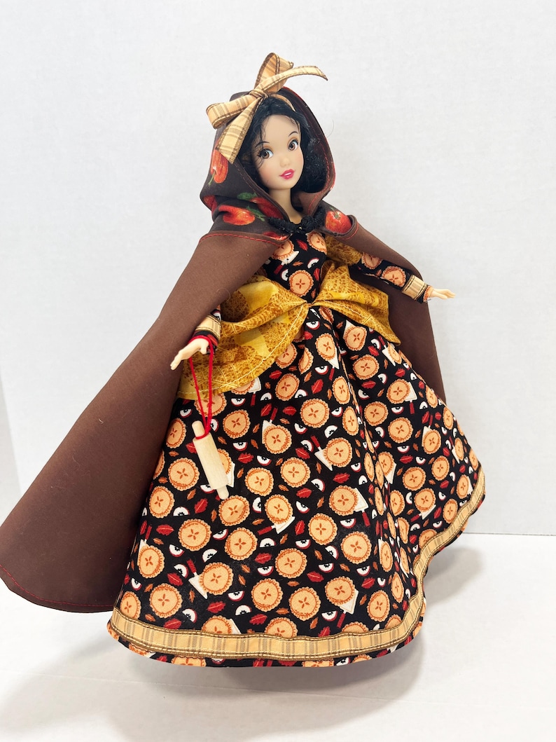 Op de afbeelding: Een pop met zwart haar draagt een bruine cape met capuchon en een geruite strik, een geel getextureerd accent in de taille en een zwarte jurk met een herhalend patroon van kleine appeltaartjes en rode appelschijfjes, met een miniatuur houten deegroller.