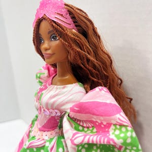 May include: A doll with brown skin and long, braided auburn hair. She wears a pink tiara and a dress with a pink, white, and green floral pattern. The dress has puffy sleeves and is embellished with sequins.