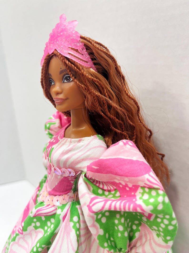 May include: A doll with brown skin and long, braided auburn hair. She wears a pink tiara and a dress with a pink, white, and green floral pattern. The dress has puffy sleeves and is embellished with sequins.