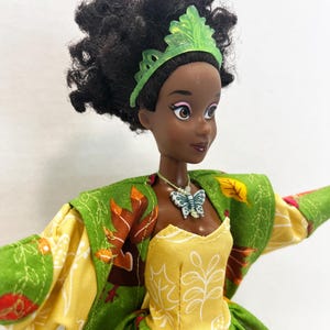K&ouml;nnte beinhalten: Eine Puppe mit dunklem, lockigem Haar tr&auml;gt ein gr&uuml;nes Diadem, ein gelbes Oberteil mit wei&szlig;en Blattumrissen und eine gr&uuml;ne Jacke mit einem Muster aus braunen und orangefarbenen Herbstbl&auml;ttern. Die Puppe tr&auml;gt einen wei&szlig;en Schmetterlingsanh&auml;nger.