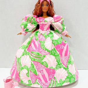 May include: A doll wearing a pink crown and a long gown with a green and white fish and seashell pattern. The doll has long brown hair and is holding a small pink watering can. The dress has puffy sleeves and a pink bodice.