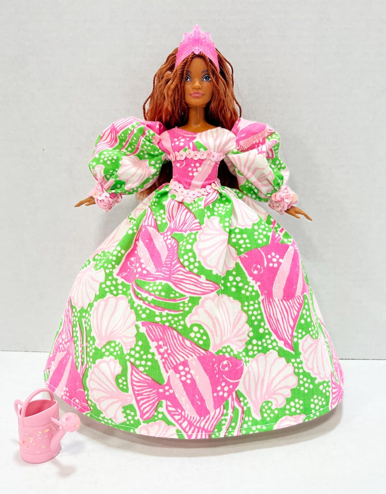 May include: A doll wearing a pink crown and a long gown with a green and white fish and seashell pattern. The doll has long brown hair and is holding a small pink watering can. The dress has puffy sleeves and a pink bodice.