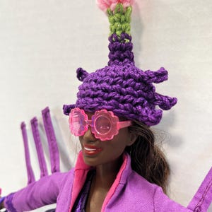 May include: A doll wearing a purple crochet hat with a pink flower and green stem, and pink flower-shaped sunglasses. The doll is also wearing a purple jacket with pink trim.