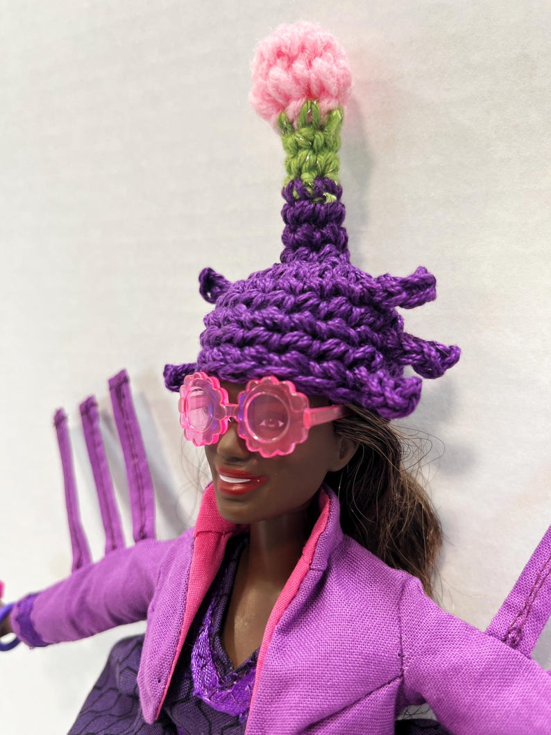 May include: A doll wearing a purple crochet hat with a pink flower and green stem, and pink flower-shaped sunglasses. The doll is also wearing a purple jacket with pink trim.