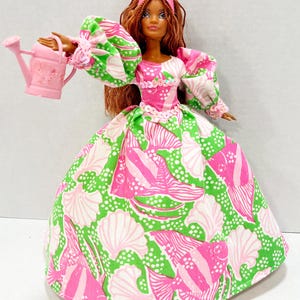 May include: A doll wearing a pink and green patterned gown, a pink crown, and holding a pink watering can. The doll has brown hair and a fair complexion. The gown features a seashell and fish pattern.