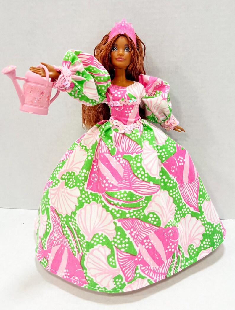 May include: A doll wearing a pink and green patterned gown, a pink crown, and holding a pink watering can. The doll has brown hair and a fair complexion. The gown features a seashell and fish pattern.