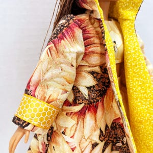 Puede incluir: Una mu&ntilde;eca con un abrigo largo con estampado de girasoles. El interior y el cuello del abrigo son de color amarillo brillante. Los pu&ntilde;os de las mangas presentan un patr&oacute;n de panal de abeja amarillo. La mu&ntilde;eca tambi&eacute;n lleva una camiseta amarilla con un gr&aacute;fico de abeja.