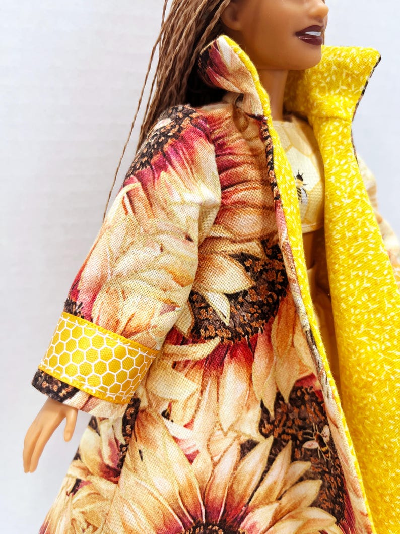 Puede incluir: Una mu&ntilde;eca con un abrigo largo con estampado de girasoles. El interior y el cuello del abrigo son de color amarillo brillante. Los pu&ntilde;os de las mangas presentan un patr&oacute;n de panal de abeja amarillo. La mu&ntilde;eca tambi&eacute;n lleva una camiseta amarilla con un gr&aacute;fico de abeja.