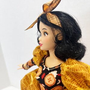 Op de afbeelding: Een pop met zwart haar in een boblijn, met een beige hoofdband met ruitjespatroon. De pop draagt een zwart lijfje met een herhalend patroon van kleine oranje en witte taartjes, gecombineerd met pofmouwen van gestructureerde goudgele stof.