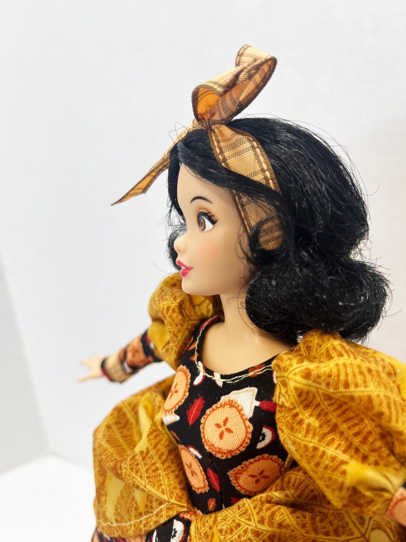 Op de afbeelding: Een pop met zwart haar in een boblijn, met een beige hoofdband met ruitjespatroon. De pop draagt een zwart lijfje met een herhalend patroon van kleine oranje en witte taartjes, gecombineerd met pofmouwen van gestructureerde goudgele stof.