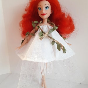 Puede incluir: Una mu&ntilde;eca de pelo rojo con un vestido blanco con un detalle de vid verde y cuerda marr&oacute;n. El vestido tiene una falda de tul y un ribete de flecos blancos.