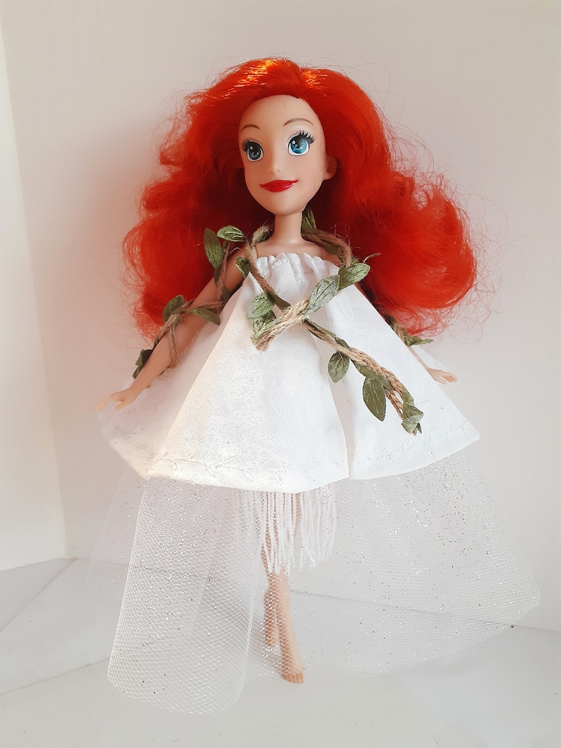 Puede incluir: Una mu&ntilde;eca de pelo rojo con un vestido blanco con un detalle de vid verde y cuerda marr&oacute;n. El vestido tiene una falda de tul y un ribete de flecos blancos.