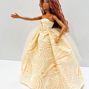 Puede incluir: Mu&ntilde;eca con pelo largo color casta&ntilde;o rojizo y una corona amarilla. La mu&ntilde;eca lleva un vestido crema sin tirantes con un estampado de conchas marinas y una enagua de tul. El brazo de la mu&ntilde;eca est&aacute; extendido.