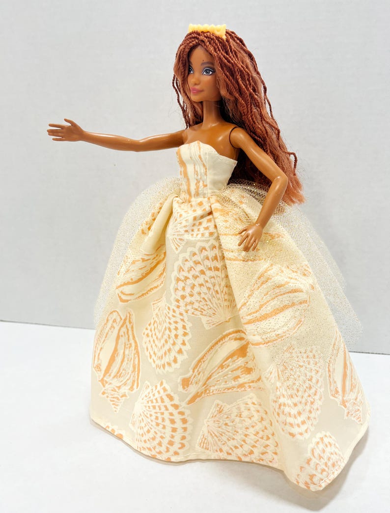Puede incluir: Mu&ntilde;eca con pelo largo color casta&ntilde;o rojizo y una corona amarilla. La mu&ntilde;eca lleva un vestido crema sin tirantes con un estampado de conchas marinas y una enagua de tul. El brazo de la mu&ntilde;eca est&aacute; extendido.