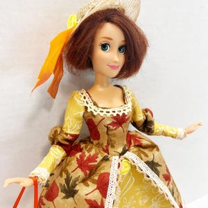 May include: A doll with shoulder-length brown hair wearing a straw hat with an orange ribbon and a dress featuring a pattern of red, brown, and yellow autumn leaves. The dress has a cream-colored lace trim at the neckline and waist.