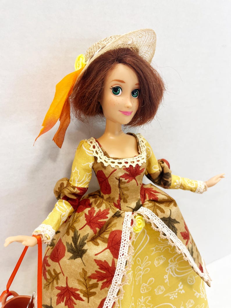 May include: A doll with shoulder-length brown hair wearing a straw hat with an orange ribbon and a dress featuring a pattern of red, brown, and yellow autumn leaves. The dress has a cream-colored lace trim at the neckline and waist.