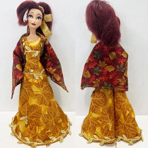 May include: A doll with dark red hair styled in a high ponytail, wearing a floor-length yellow dress featuring a brown leaf pattern and a gold leaf trim at the hem. The doll wears a matching cape with a multi-colored autumn leaf print and gold accents.