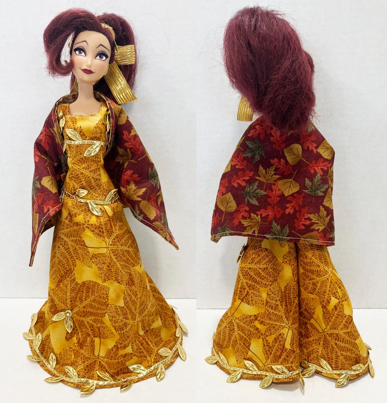 May include: A doll with dark red hair styled in a high ponytail, wearing a floor-length yellow dress featuring a brown leaf pattern and a gold leaf trim at the hem. The doll wears a matching cape with a multi-colored autumn leaf print and gold accents.