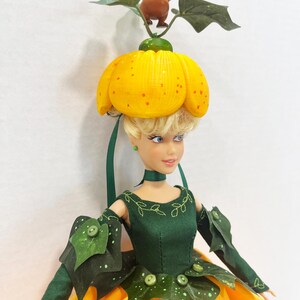 May include: A doll with blonde hair wearing a yellow pumpkin-shaped hat topped with a small brown mouse figure and green leaves. The doll wears a dark green bodice with leaf-shaped accents and buttons, paired with a bright yellow skirt featuring white circular embroidery details.