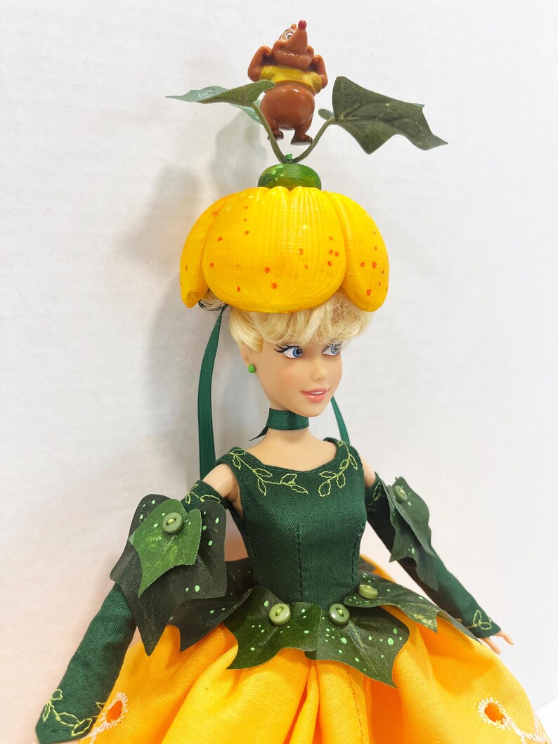 May include: A doll with blonde hair wearing a yellow pumpkin-shaped hat topped with a small brown mouse figure and green leaves. The doll wears a dark green bodice with leaf-shaped accents and buttons, paired with a bright yellow skirt featuring white circular embroidery details.