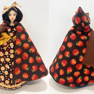 Op de afbeelding: Een pop in een jurk met taartpatroon en een bruine cape met een rode appelprint. De pop heeft donker haar, een bruine strik en houdt een kleine witte deegroller vast. De cape is van voor en achter te zien met de bedrukte appelstof.