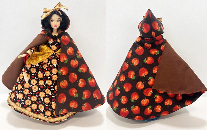 Op de afbeelding: Een pop in een jurk met taartpatroon en een bruine cape met een rode appelprint. De pop heeft donker haar, een bruine strik en houdt een kleine witte deegroller vast. De cape is van voor en achter te zien met de bedrukte appelstof.