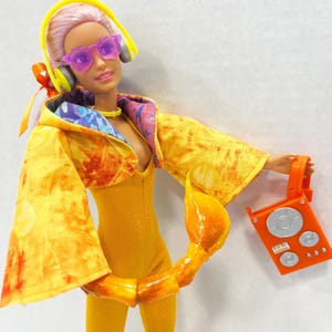 K&ouml;nnte beinhalten: Eine Puppe mit rosa Haaren tr&auml;gt eine violette Sonnenbrille, gelbe Kopfh&ouml;rer und einen gelben Overall unter einer gelb-orangefarbenen Kapuzenjacke im Batik-Look. Ein gelber Skorpionschwanz ist an der Taille befestigt und h&auml;lt ein orangefarbenes Radio.
