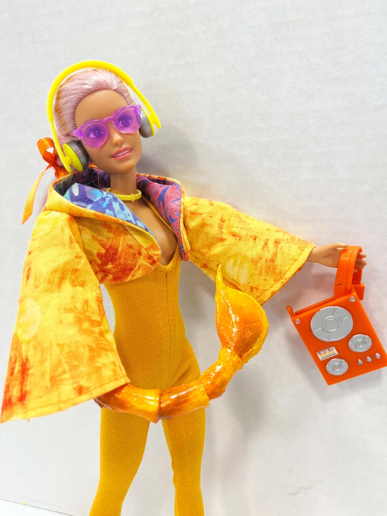 K&ouml;nnte beinhalten: Eine Puppe mit rosa Haaren tr&auml;gt eine violette Sonnenbrille, gelbe Kopfh&ouml;rer und einen gelben Overall unter einer gelb-orangefarbenen Kapuzenjacke im Batik-Look. Ein gelber Skorpionschwanz ist an der Taille befestigt und h&auml;lt ein orangefarbenes Radio.