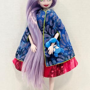 Pu&ograve; includere: Una bambola con lunghi capelli color lavanda, che indossa un mantello blu con un motivo a foglie scure e bordino giallo neon sopra una gonna rossa. La bambola tiene in mano una piccola figura di granchio blu e indossa sandali viola con tacco.