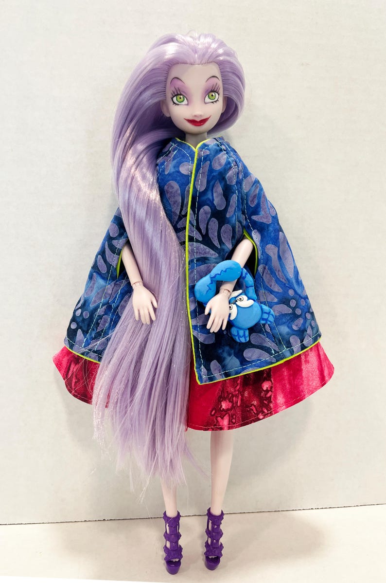Pu&ograve; includere: Una bambola con lunghi capelli color lavanda, che indossa un mantello blu con un motivo a foglie scure e bordino giallo neon sopra una gonna rossa. La bambola tiene in mano una piccola figura di granchio blu e indossa sandali viola con tacco.