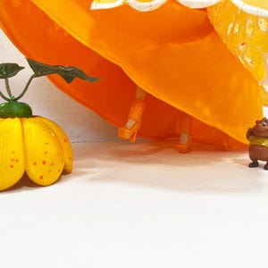May include: A doll wearing a bright orange dress with a white scalloped eyelet hem, orange platform heels, and orange legs. Beside the doll is a yellow pumpkin-shaped object with green leaves and a small brown cartoon character figure.