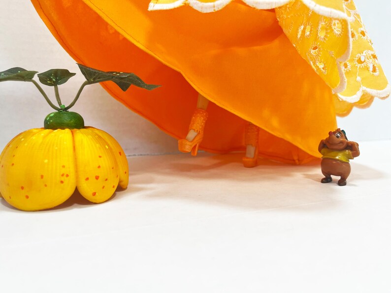 May include: A doll wearing a bright orange dress with a white scalloped eyelet hem, orange platform heels, and orange legs. Beside the doll is a yellow pumpkin-shaped object with green leaves and a small brown cartoon character figure.