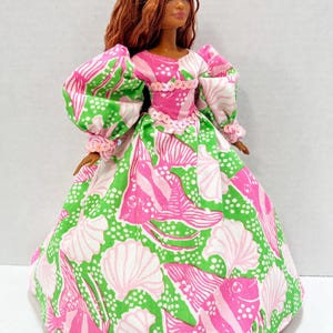 May include: A doll wearing a pink and green patterned dress with a matching pink tiara. The dress features a seashell and fish design. The doll has brown hair and is wearing a pink tiara. The dress has puffy sleeves and a full skirt.